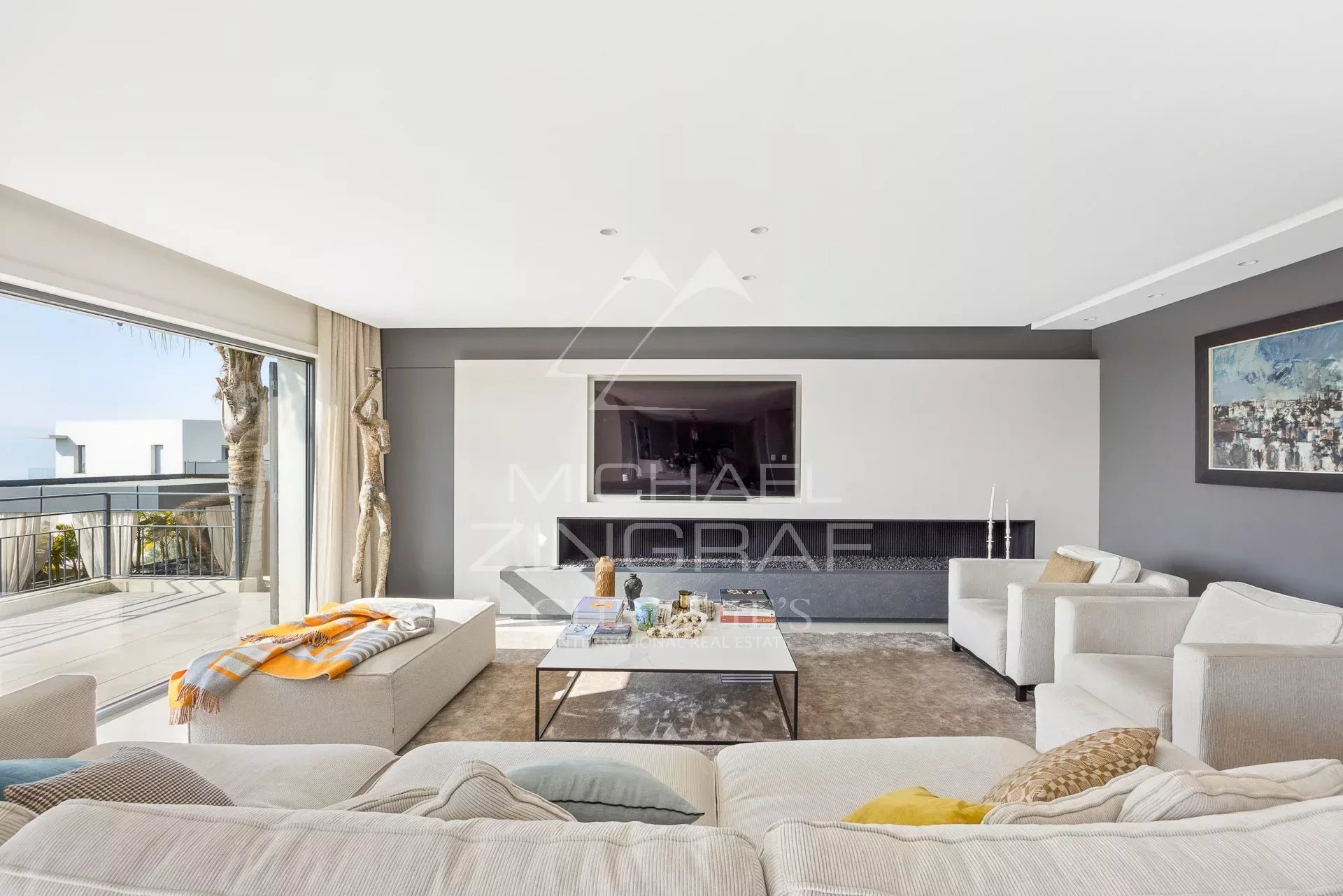 Bright modern living room with white sofas, a low glass coffee table, gray walls, and a balcony with an ocean view.