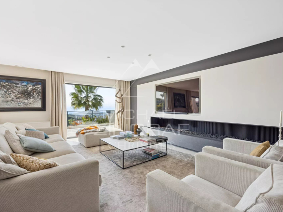 Bright contemporary living room with beige sofa set, glass coffee table, and a long fireplace, opening to a balcony with an ocean view and palm trees.