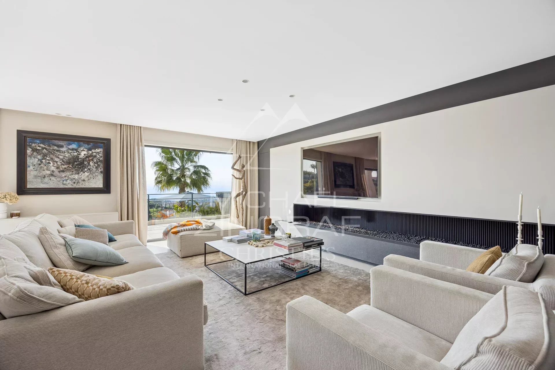 Bright contemporary living room with beige sofa set, glass coffee table, and a long fireplace, opening to a balcony with an ocean view and palm trees.