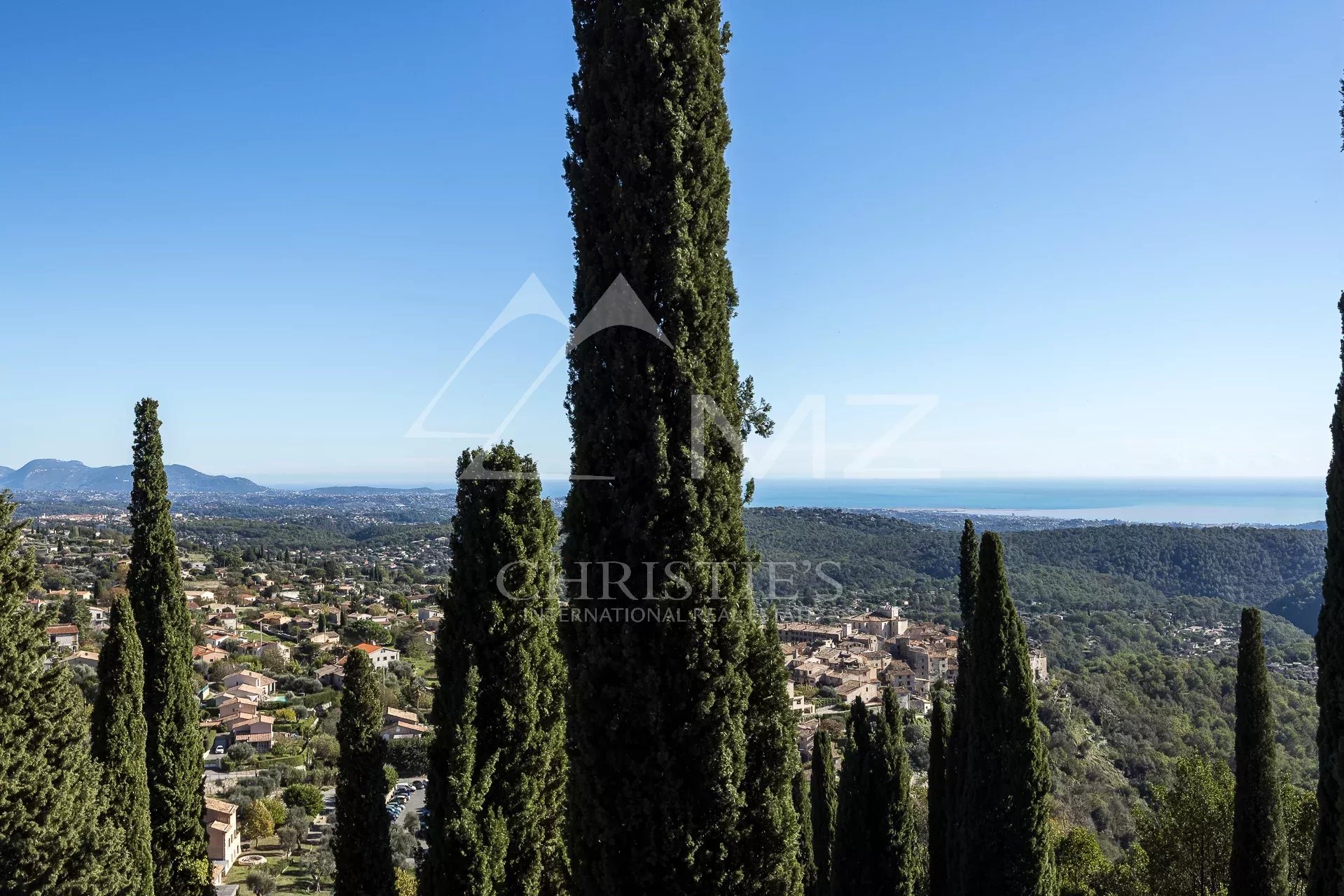 Panoramic view of Tourrettes-sur-Loup and the sea from a property with cypress trees.