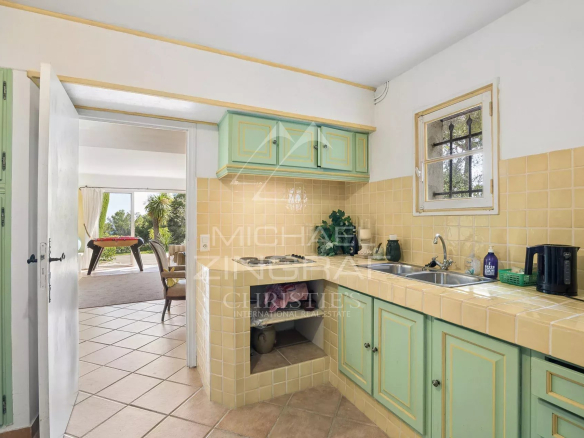 Kitchen of a villa with beige tiles and green furniture, overlooking a bright living space.