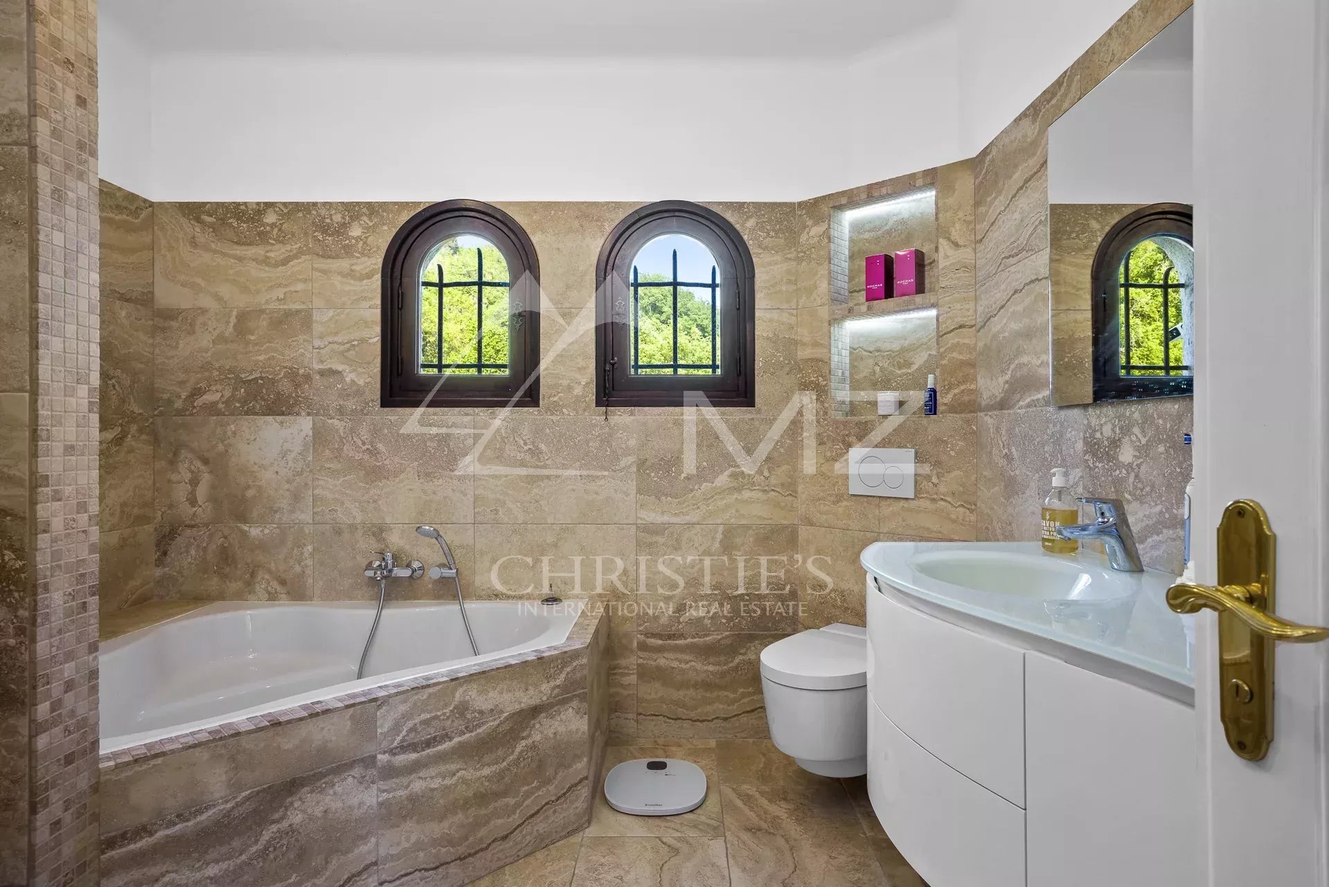 Modern bathroom with bathtub and sink, beige stone walls, arched windows.