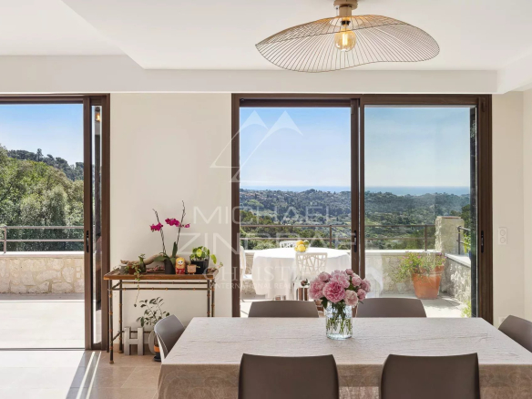 Intérieur d'une villa moderne avec vue sur la mer et le village, décoré avec une table à manger et des fenêtres ouvertes.