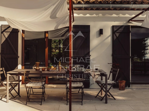 Terrace of a renovated property with wooden furniture and chairs, surrounded by greenery.