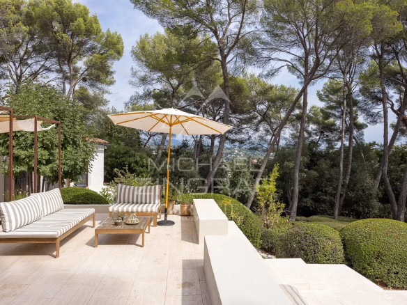 Terrace of a renovated property with garden furniture and parasol, surrounded by trees in a landscaped park.