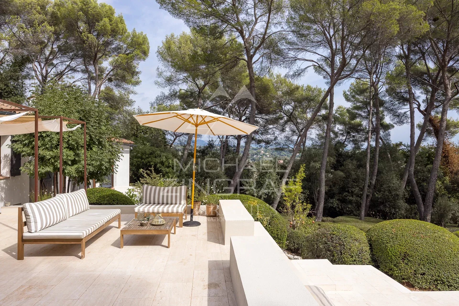 Terrace of a renovated villa in La Colle-sur-Loup Terrace of a renovated property with garden furniture and parasol, surrounded by trees in a landscaped park.