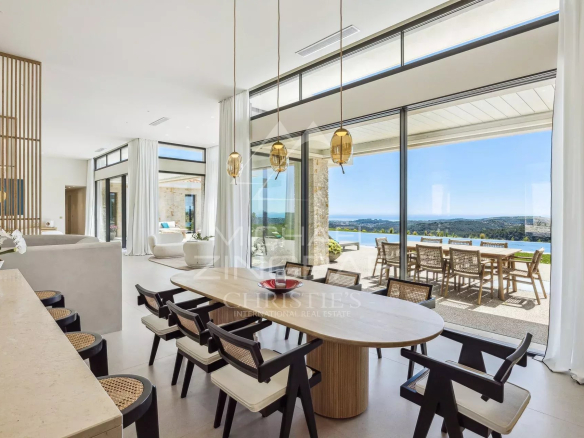 Open-plan dining area with a round wooden table and black-and-white chairs, glass walls, and an ocean-view terrace.