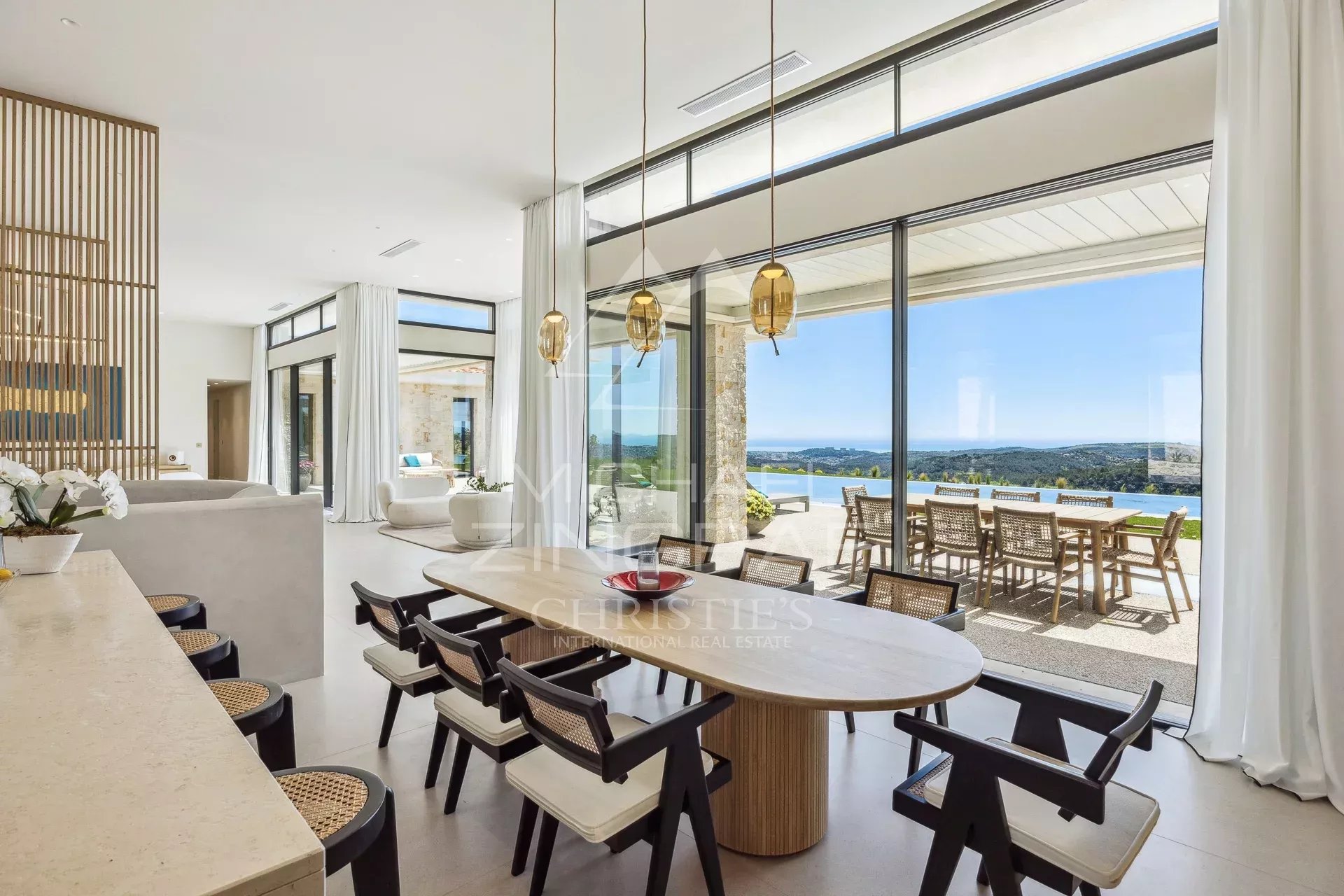 Open-plan dining area with a round wooden table and black-and-white chairs, glass walls, and an ocean-view terrace.