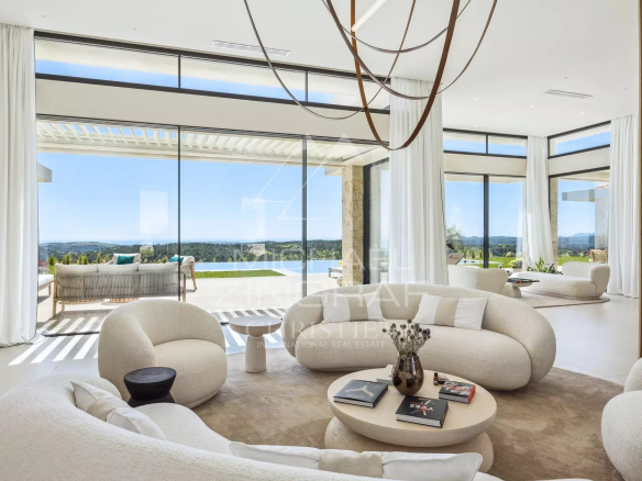 Bright modern living room with white curved sofas, a round coffee table, and floor-to-ceiling glass walls overlooking a pool and landscape outside.