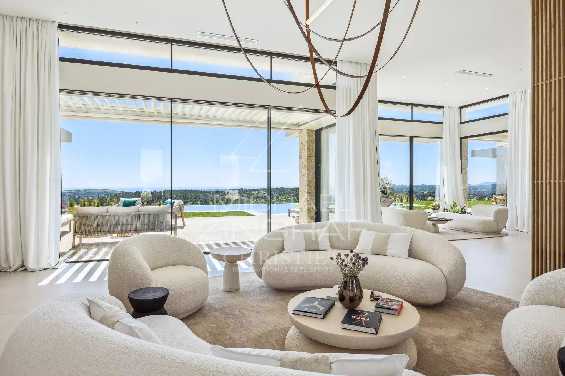 Bright modern living room with white curved sofas, a round coffee table, and floor-to-ceiling glass walls overlooking a pool and landscape outside.