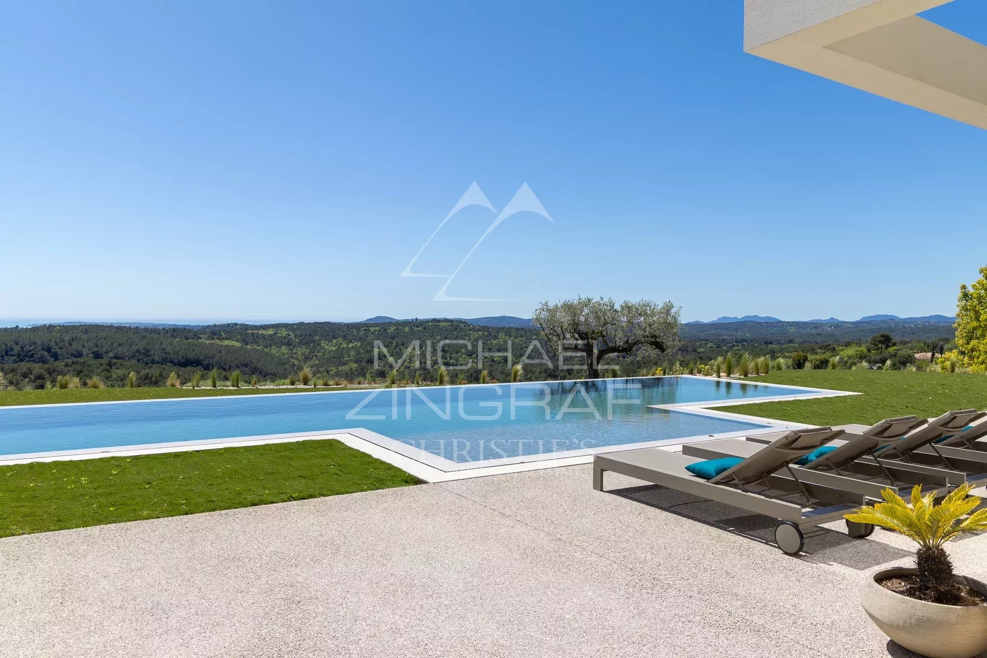 Infinity pool with white coping and lounge chairs, overlooking rolling green hills under a clear blue sky at a luxury outdoor space.