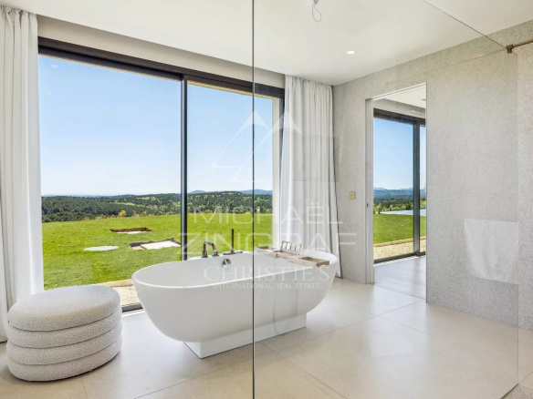 Bright, modern bathroom with a freestanding white tub by floor-to-ceiling windows overlooking a green landscape. A glass divider and light gray walls frame the scene.