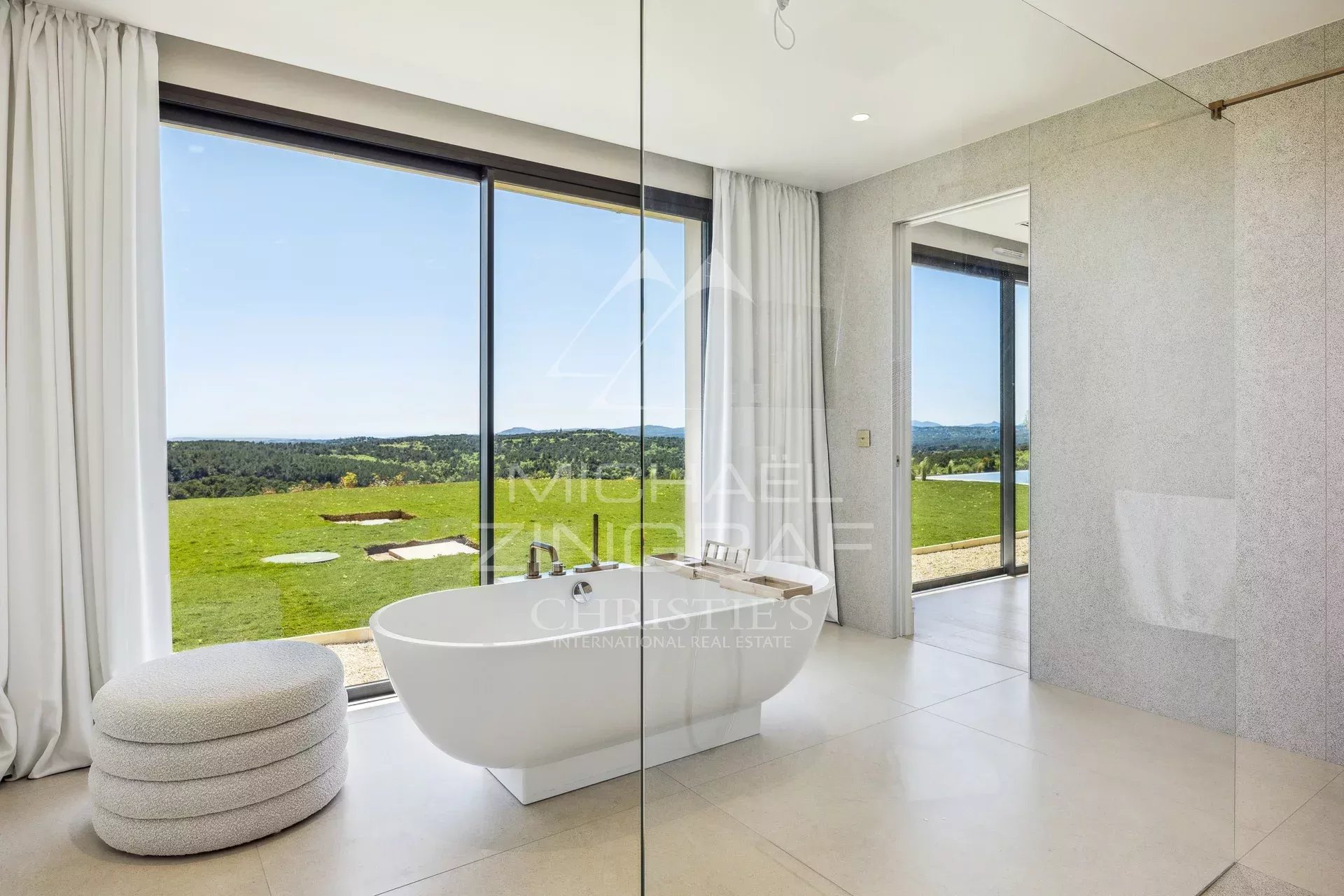 Bright, modern bathroom with a freestanding white tub by floor-to-ceiling windows overlooking a green landscape. A glass divider and light gray walls frame the scene.