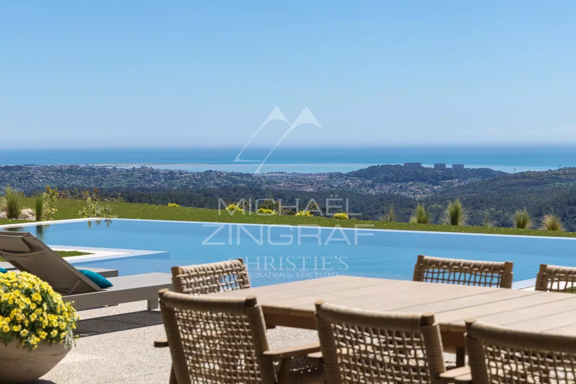 Infinity pool with wooden table and wicker chairs, overlooking rolling hills and the sea on a sunny day.