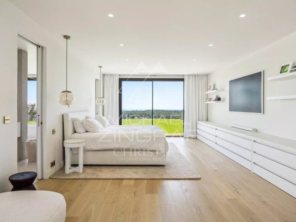 Minimalist white bedroom with a low bed, glass pendant lights, and a long white dresser, opening to a green landscape view through a large sliding door spaciously lit by natural light.