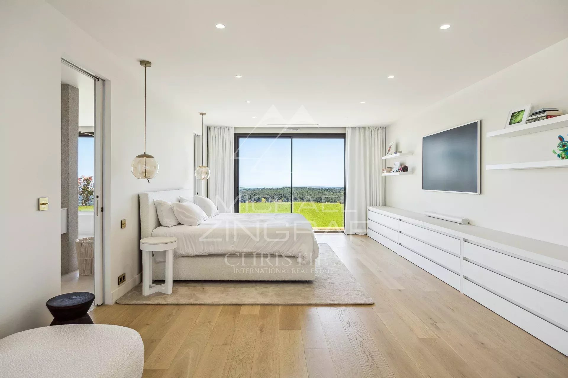 Minimalist white bedroom with a low bed, glass pendant lights, and a long white dresser, opening to a green landscape view through a large sliding door spaciously lit by natural light.