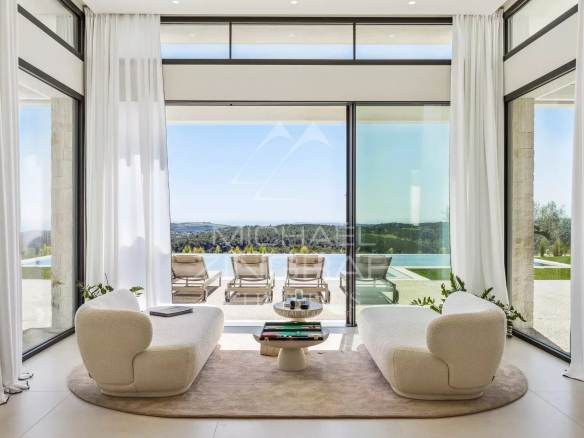 Contemporary living room with two curved white sofas on a circular rug facing a glass wall, opening to an infinity pool and scenic landscape beyond.