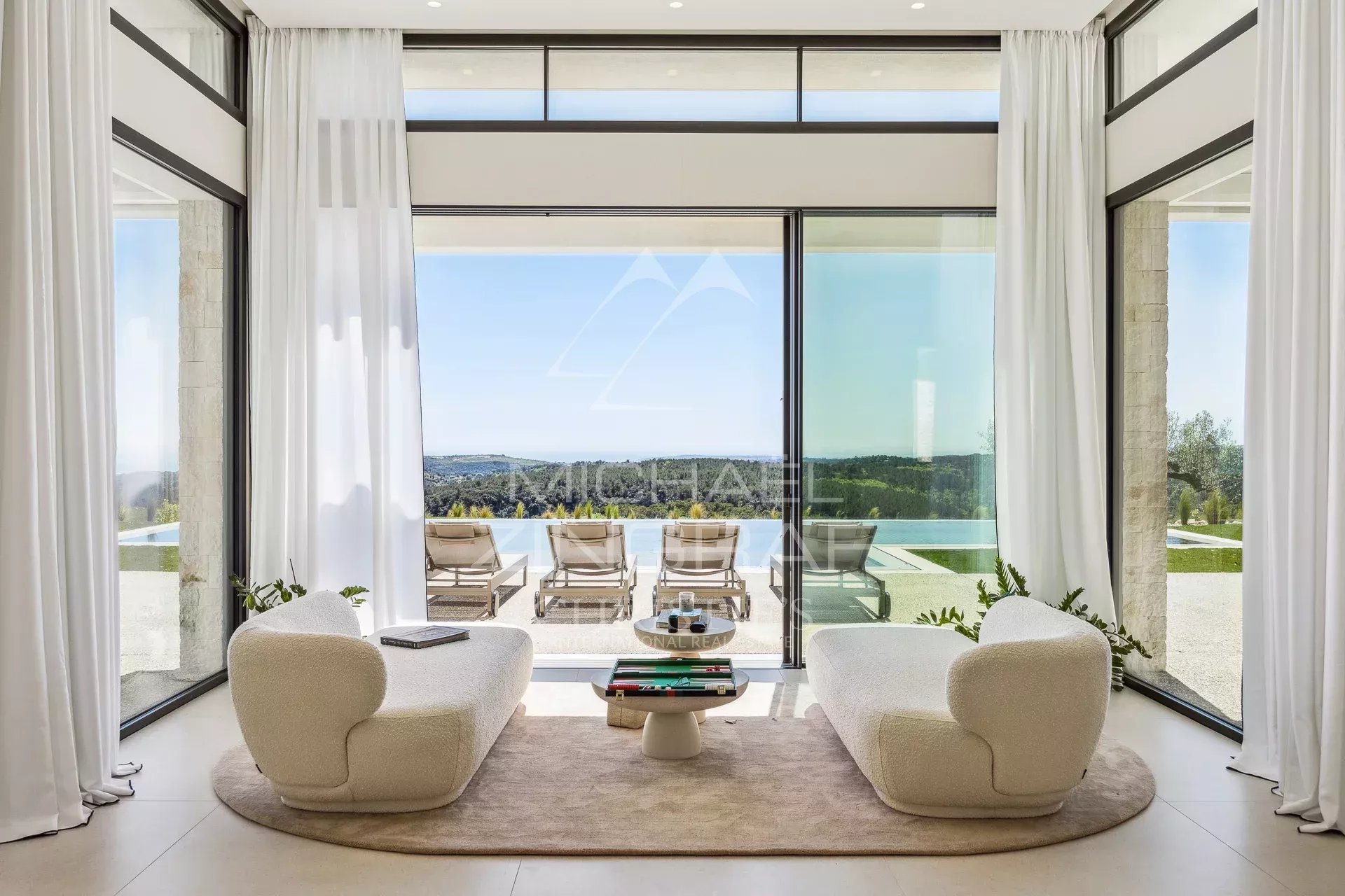Contemporary living room with two curved white sofas on a circular rug facing a glass wall, opening to an infinity pool and scenic landscape beyond.
