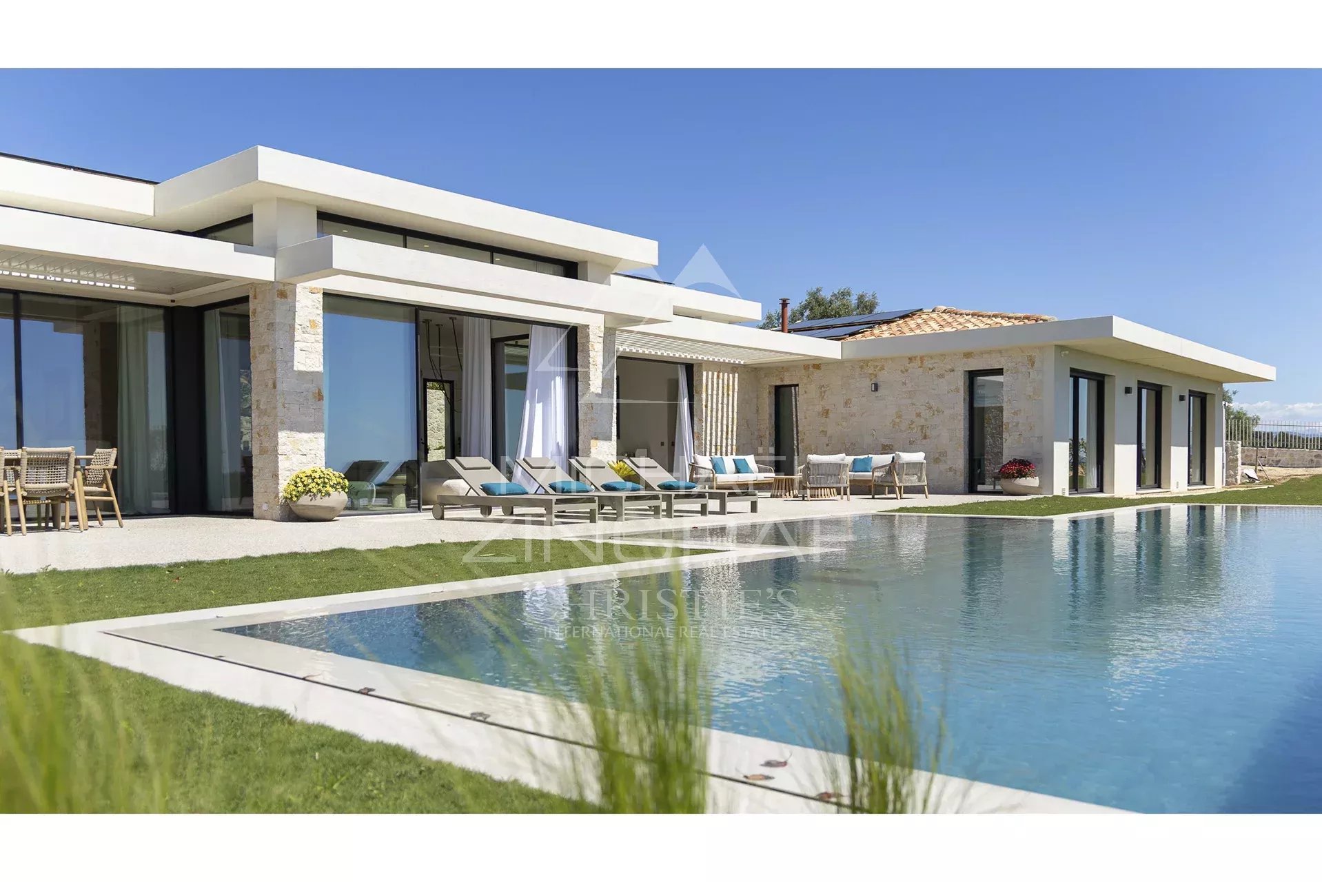Modern white villa with stone accents, a long pool, and sun loungers along a sunny patio.