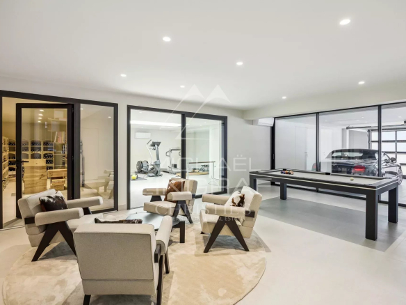 Luxurious modern basement with a circular rug seating area, glass-walled gym in the back, a billiards table, and a car in the adjacent garage area