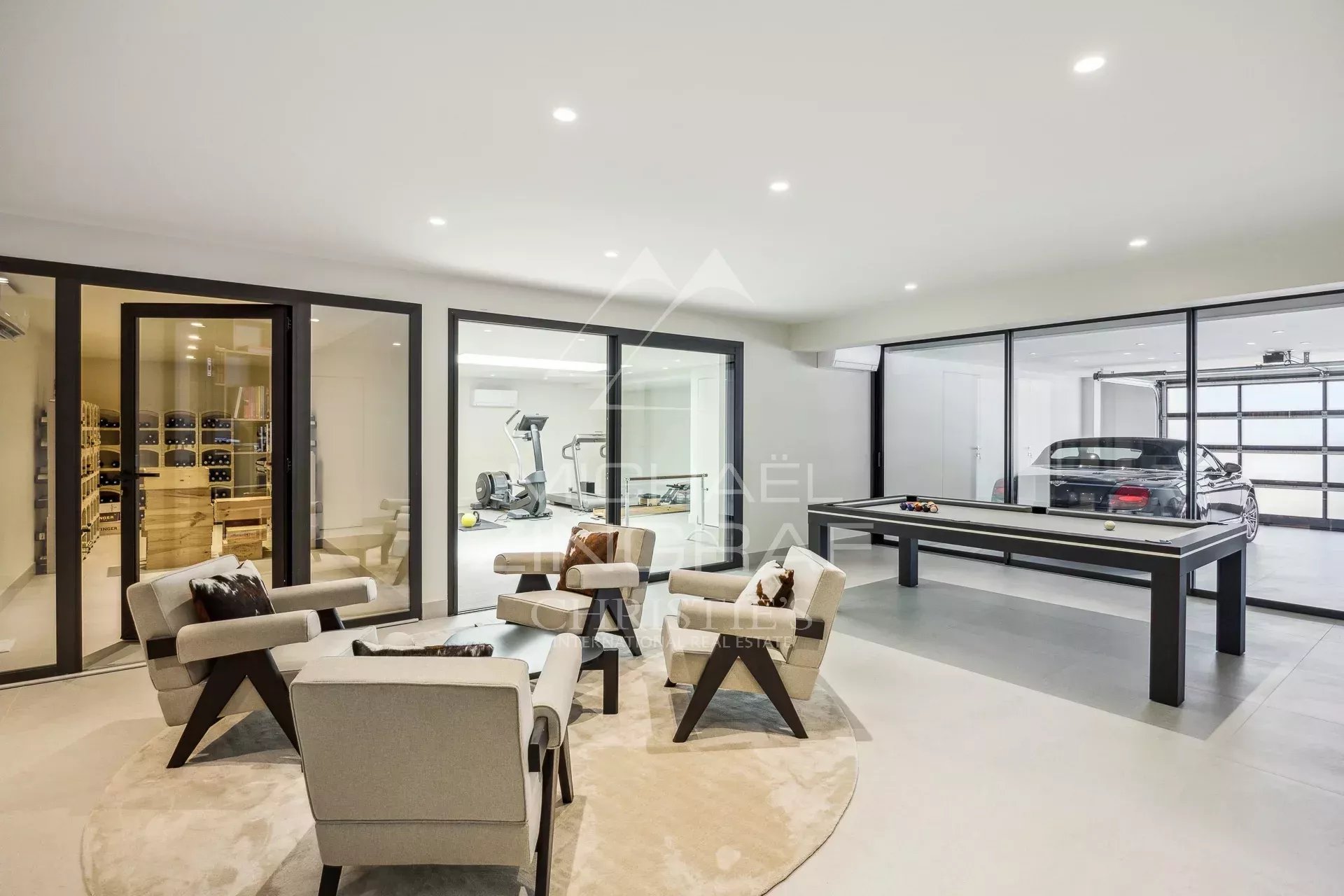 Luxurious modern basement with a circular rug seating area, glass-walled gym in the back, a billiards table, and a car in the adjacent garage area