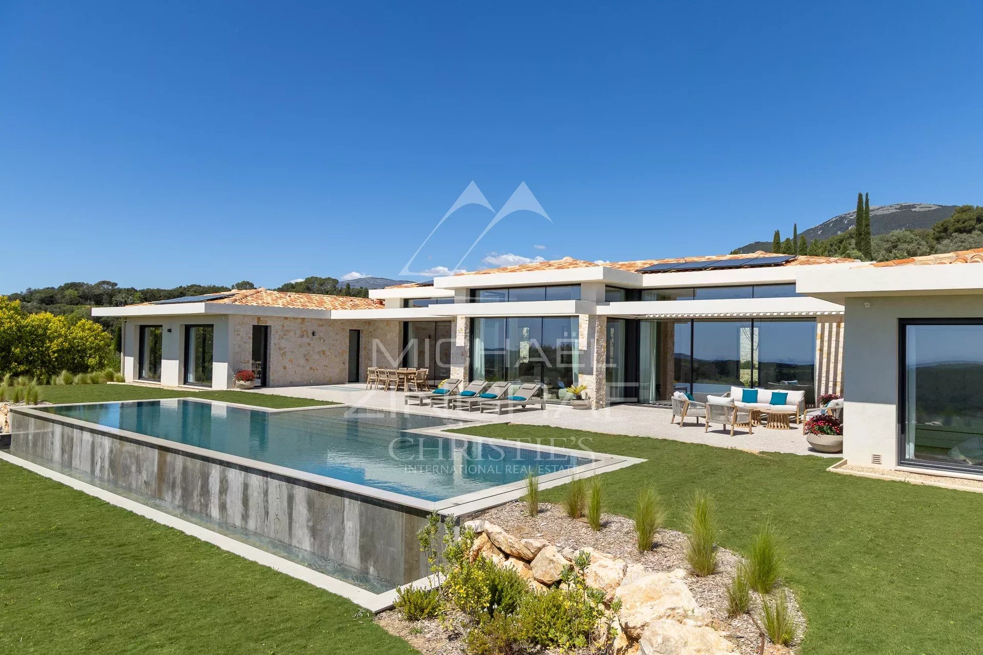 Modern single-story villa with glass walls, stone accents, and an elongated pool under a blue sky.