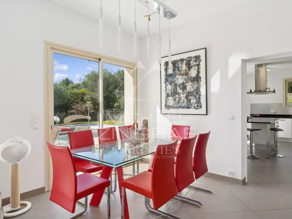 Salle à manger moderne avec table en verre et chaises rouges, vue sur un jardin verdoyant.