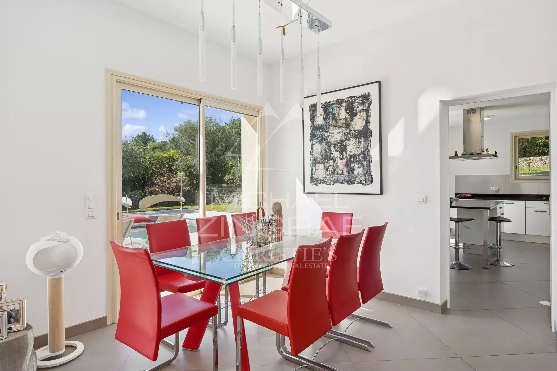 Salle à manger moderne avec table en verre et chaises rouges, vue sur un jardin verdoyant.