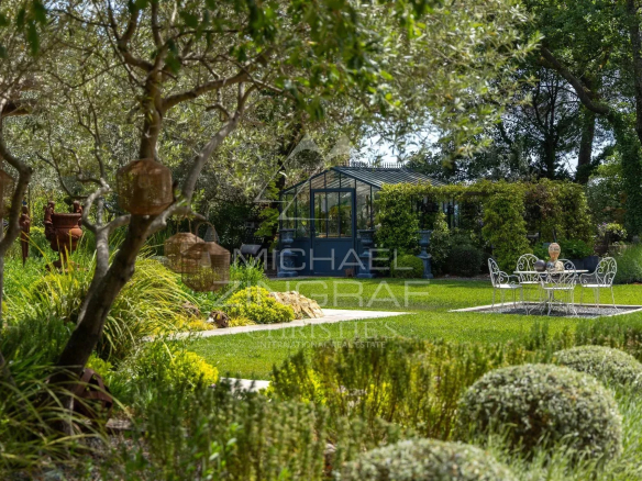 Garden of a restored estate with plants, a table and chairs, and a greenhouse in the background.