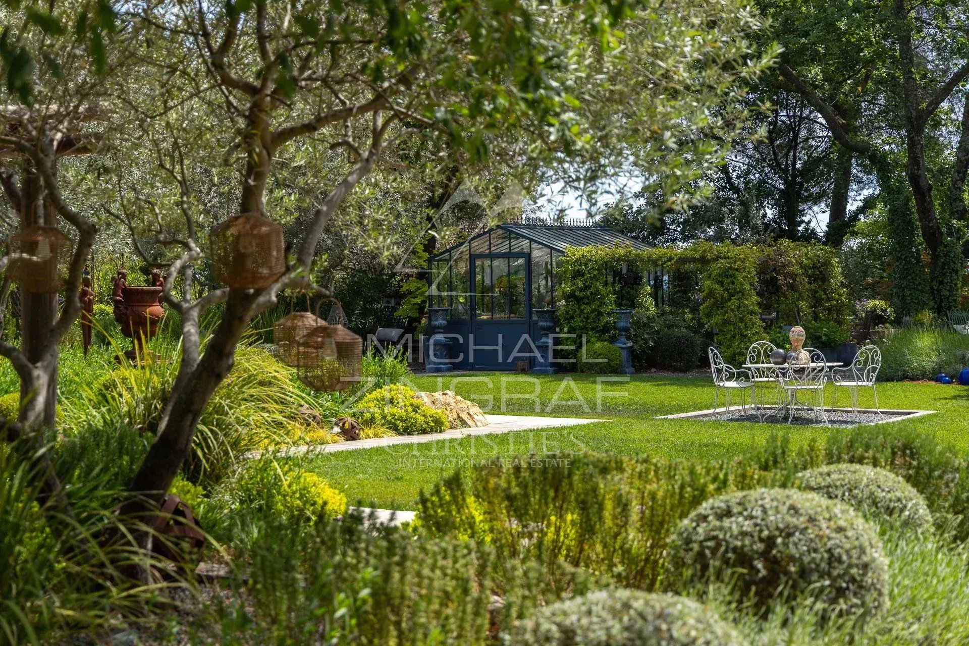 Garden of a restored estate with plants, a table and chairs, and a greenhouse in the background.