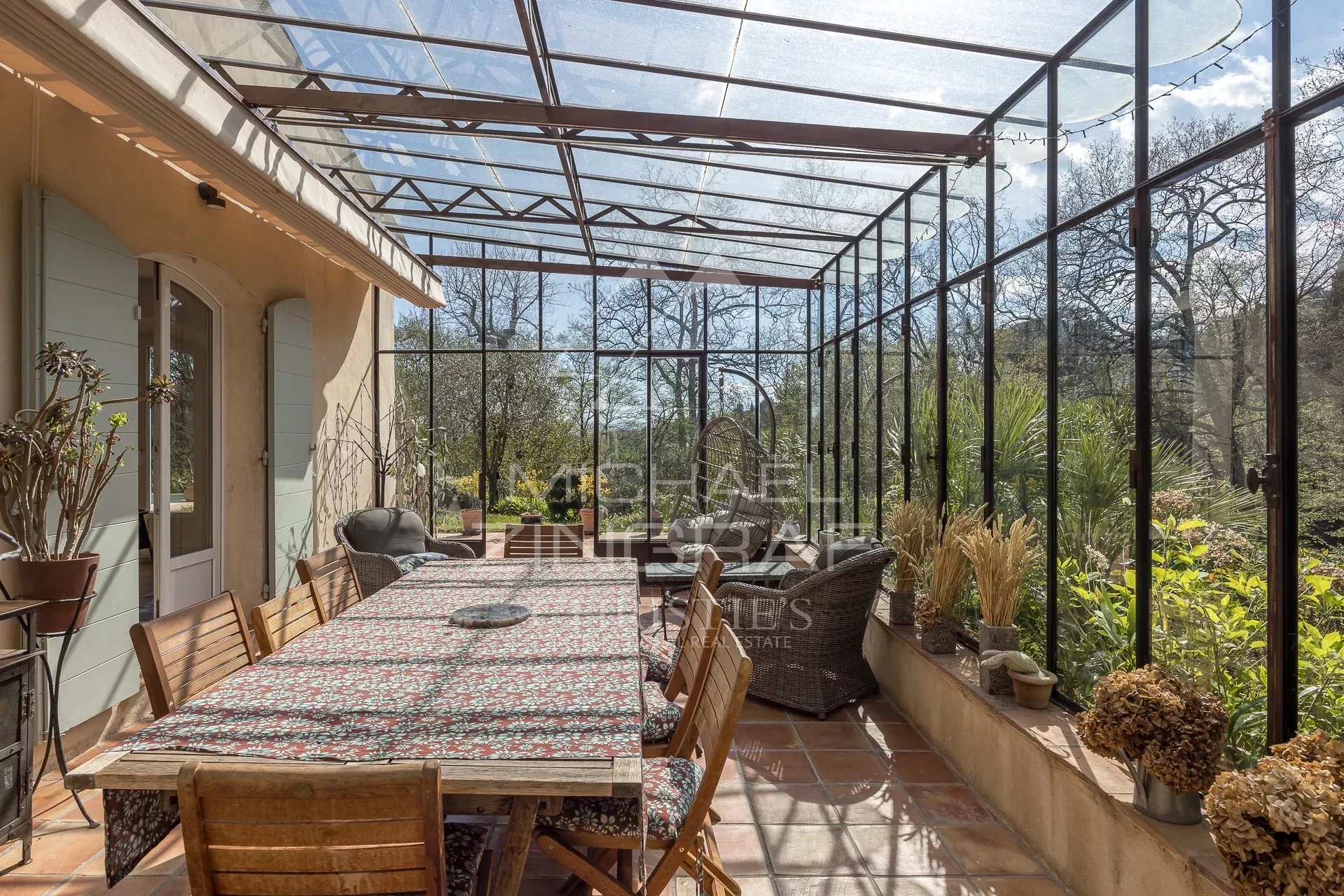 Véranda de bastide à Grasse Véranda lumineuse avec table en bois et chaises, entourée de verdure dans un parc arboré à Grasse.