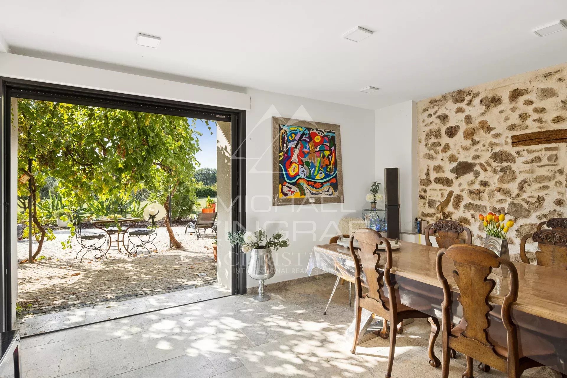 Intérieur d’une magnanerie réhabilitée Intérieur d'une demeure avec table en bois et vue sur un jardin avec des chaises en fer forgé.