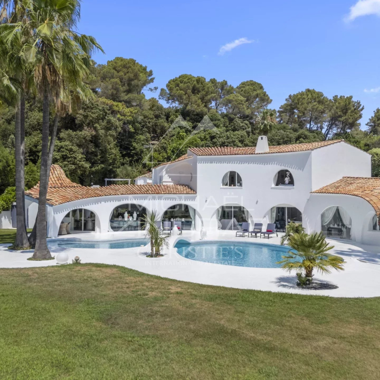 White Mediterranean-style villa with red-tiled roof and curved archways surrounding a blue swimming pool, set among palm trees and a green lawn on a sunny day.