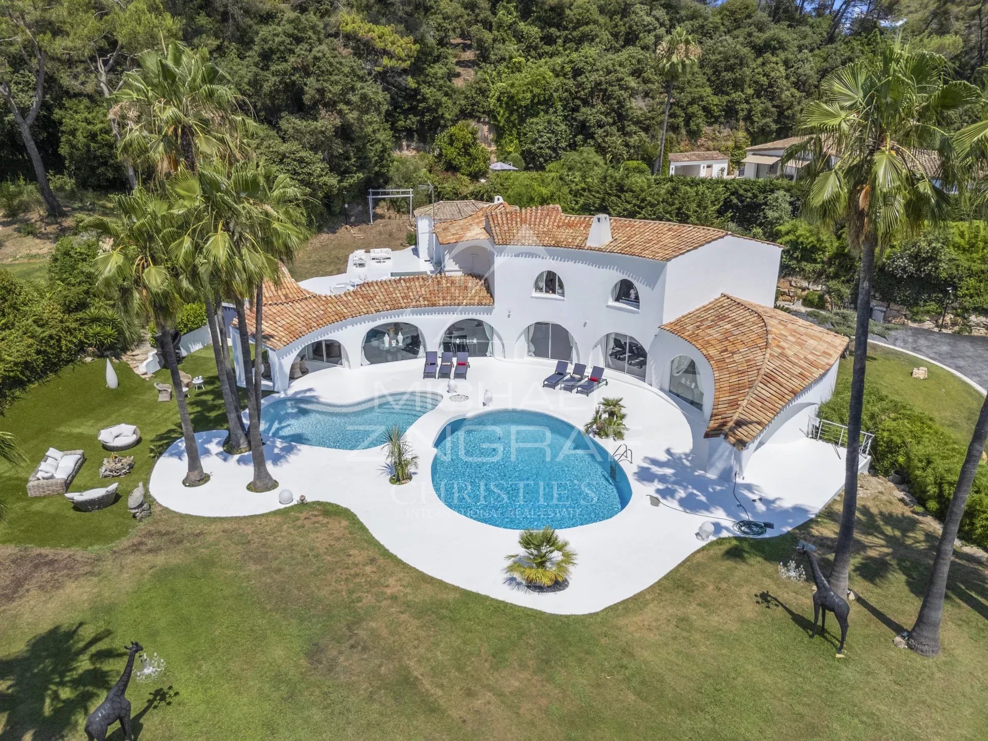 Aerial view of a white Mediterranean villa with red-tiled roofs and a curvy blue pool surrounded by palm trees and lawn.