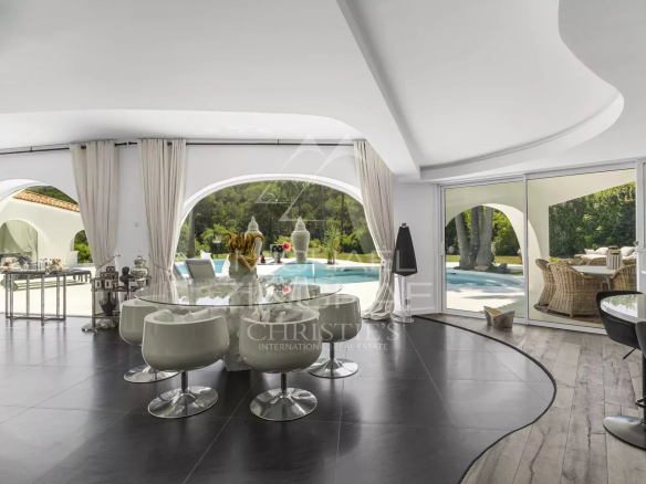 Open, modern living-dining space with curved white ceilings, glass dining table and white swivel chairs overlooking a pool and garden through arched windows.