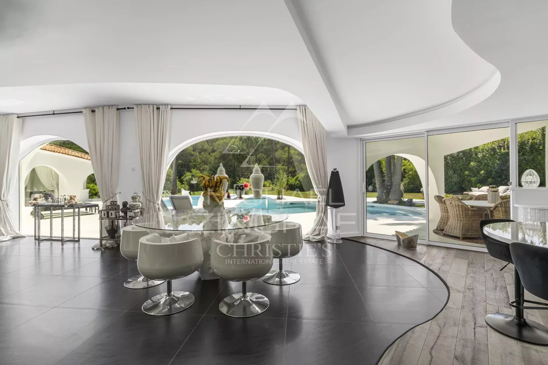 Open, modern living-dining space with curved white ceilings, glass dining table and white swivel chairs overlooking a pool and garden through arched windows.