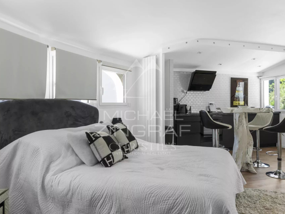 Studio apartment with a white quilted bed in the foreground and a modern kitchenette with a breakfast bar and black stools in the background.