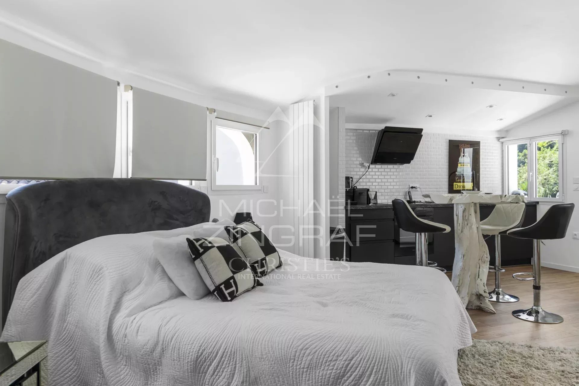 Studio apartment with a white quilted bed in the foreground and a modern kitchenette with a breakfast bar and black stools in the background.