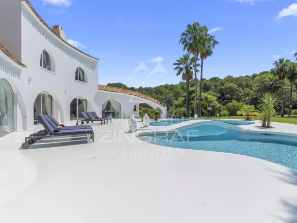 White Mediterranean villa with curved walls, arched windows, and a large swimming pool surrounded by lounge chairs and palm trees.
