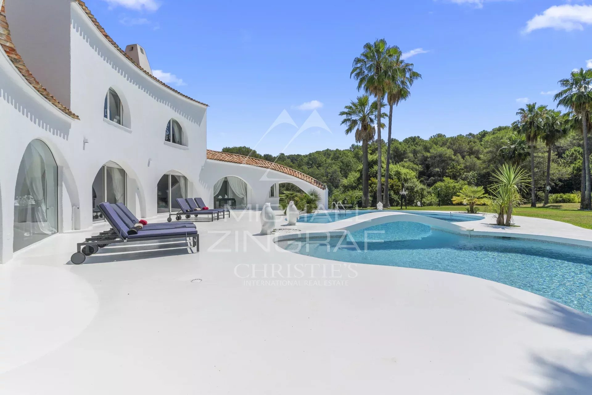 White Mediterranean villa with curved walls, arched windows, and a large swimming pool surrounded by lounge chairs and palm trees.