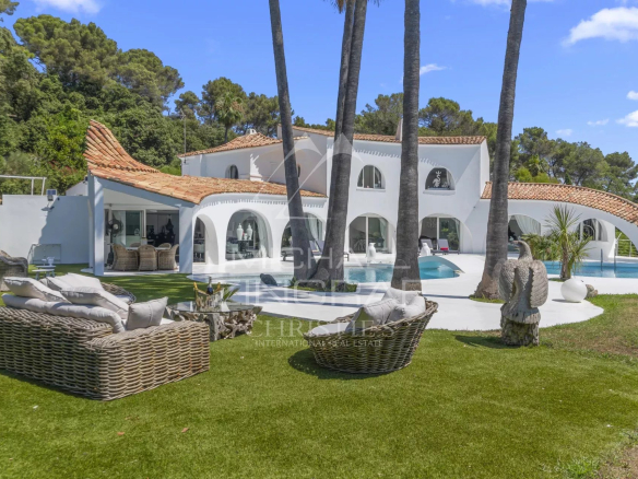White Mediterranean villa with orange-tiled roof, curved archways, and a curved poolside patio with wicker lounge seating on a green lawn.