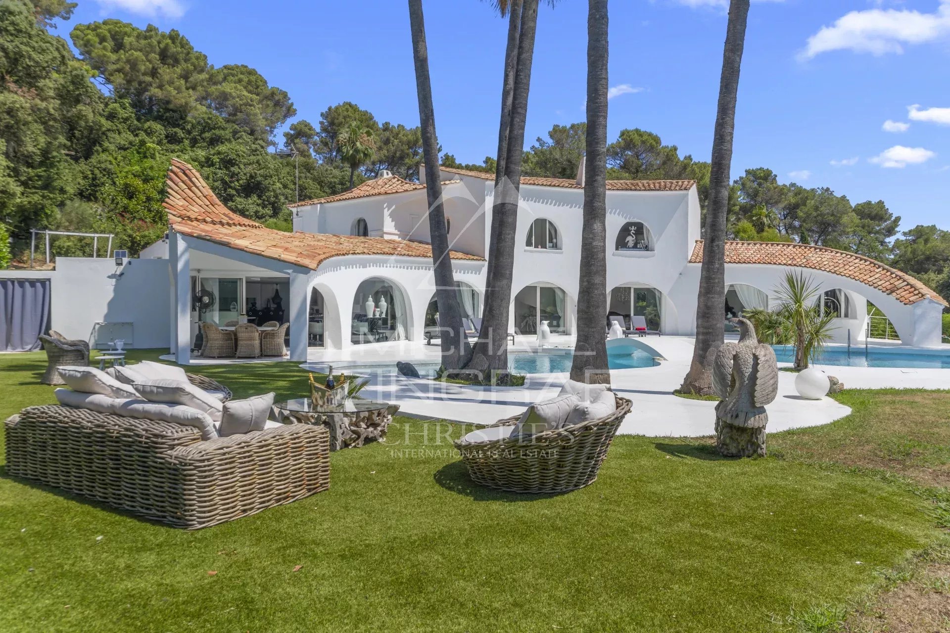 White Mediterranean villa with orange-tiled roof, curved archways, and a curved poolside patio with wicker lounge seating on a green lawn.