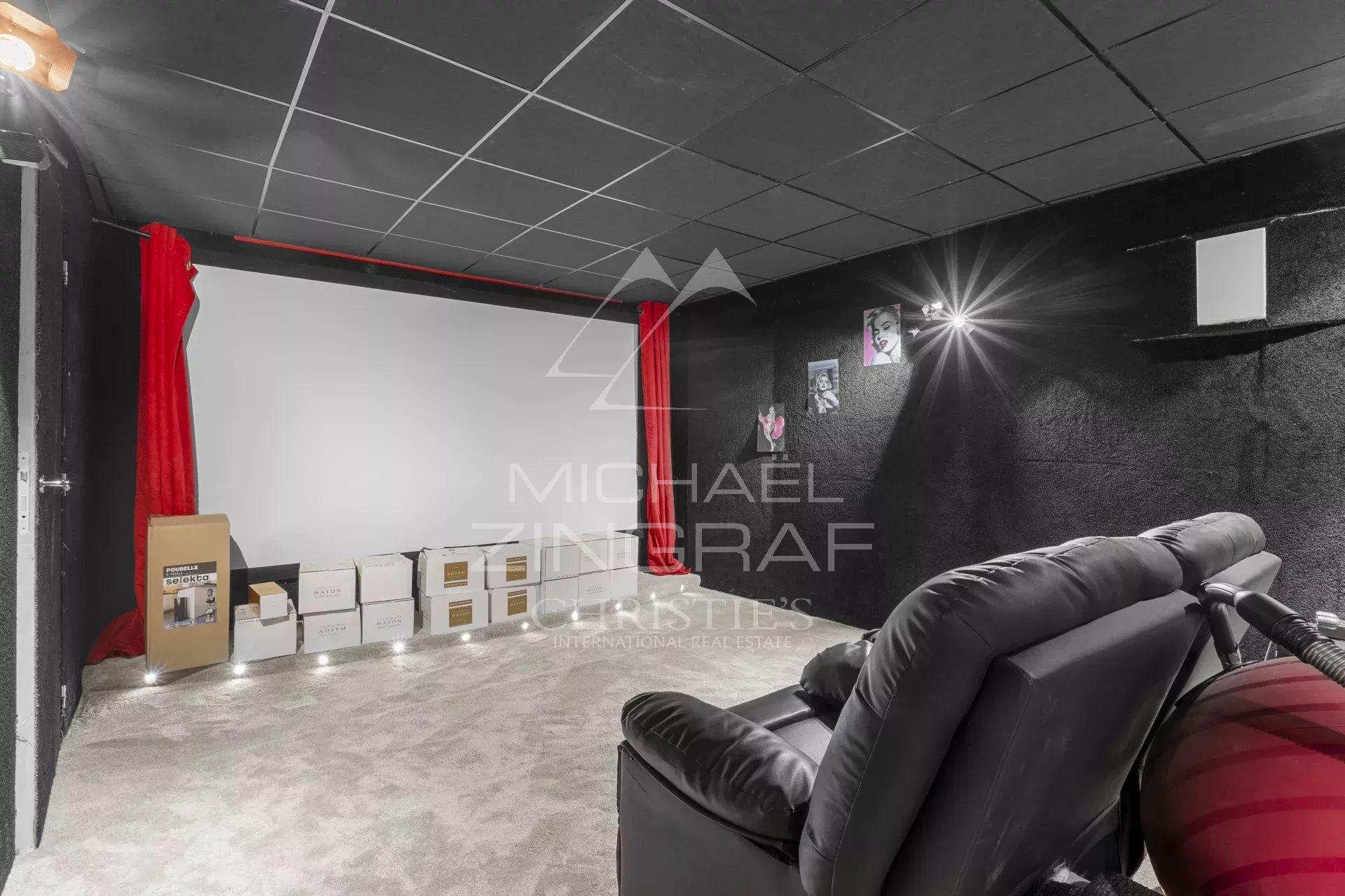 Home cinema room with a large white projection screen, red curtains, and a black-wall background; boxes line the baseboard and a plush recliner is in the foreground.