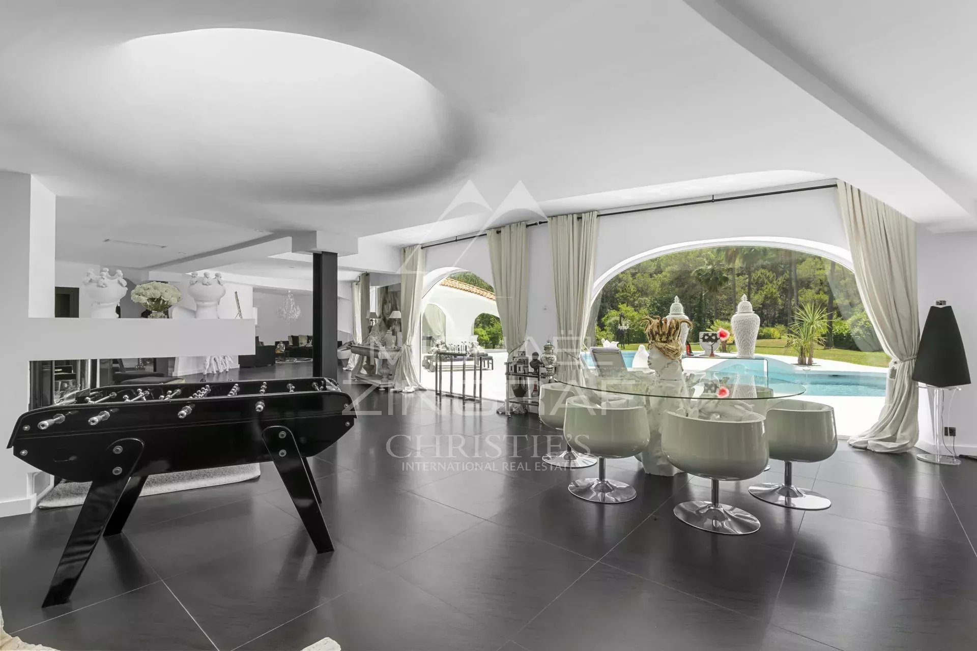 Open-plan living area with a black foosball table on dark tile, glass dining setup, and poolside garden view through large arches.