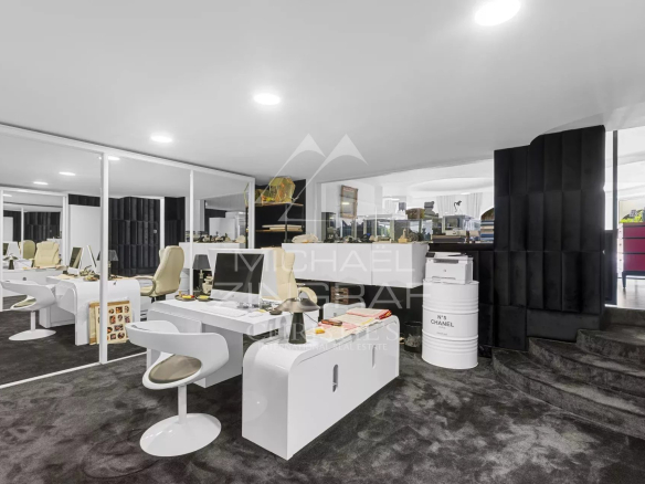 Open-plan modern showroom/office with white desks and chairs, glass partitions, a black padded wall, and a raised area with a staircase.