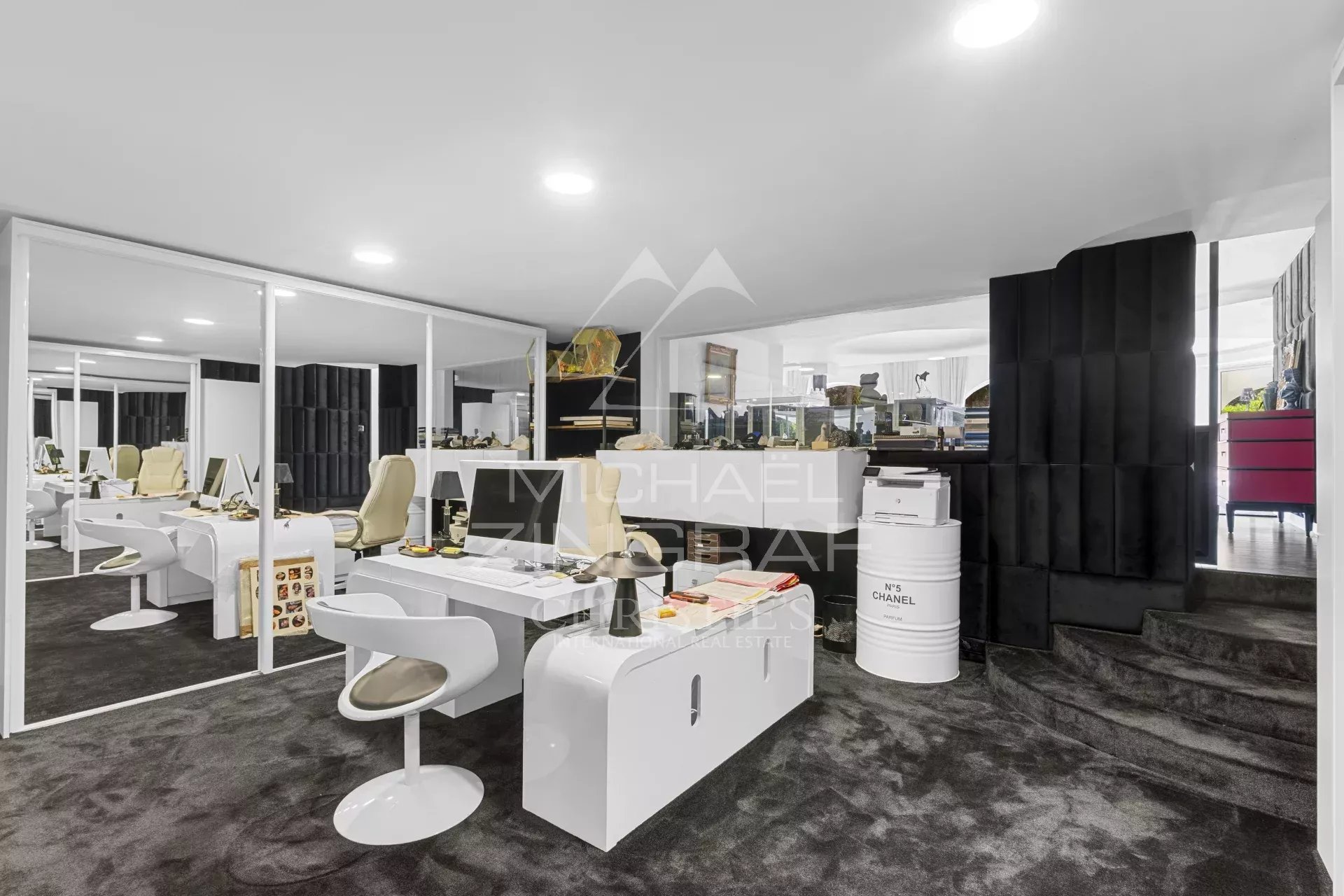 Open-plan modern showroom/office with white desks and chairs, glass partitions, a black padded wall, and a raised area with a staircase.