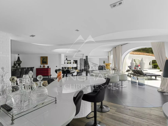 Luxurious white open-plan living area with a curved ceiling, glass bar top with crystal decanters, and arched windows opening to a poolside lounge.