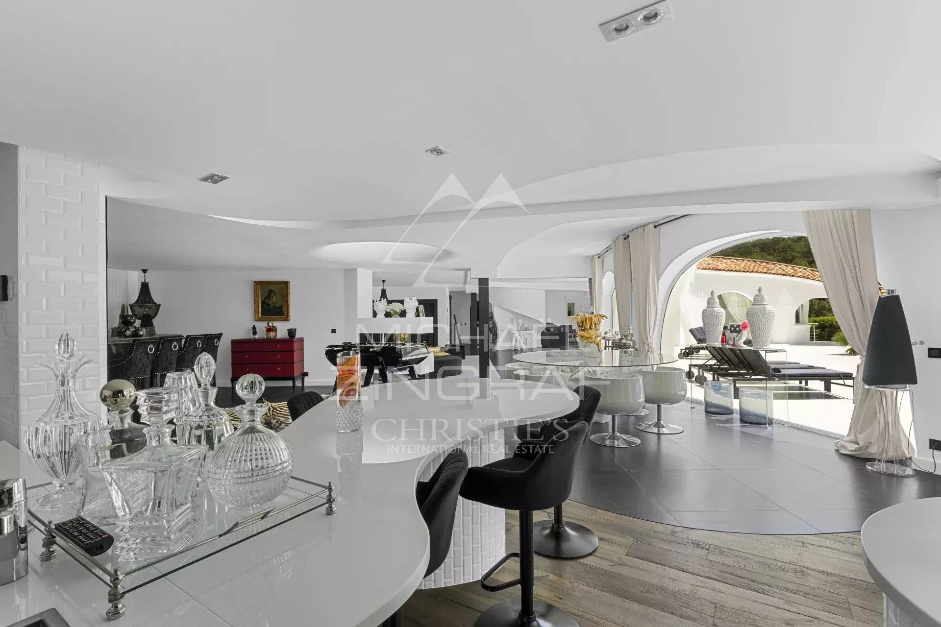 Luxurious white open-plan living area with a curved ceiling, glass bar top with crystal decanters, and arched windows opening to a poolside lounge.