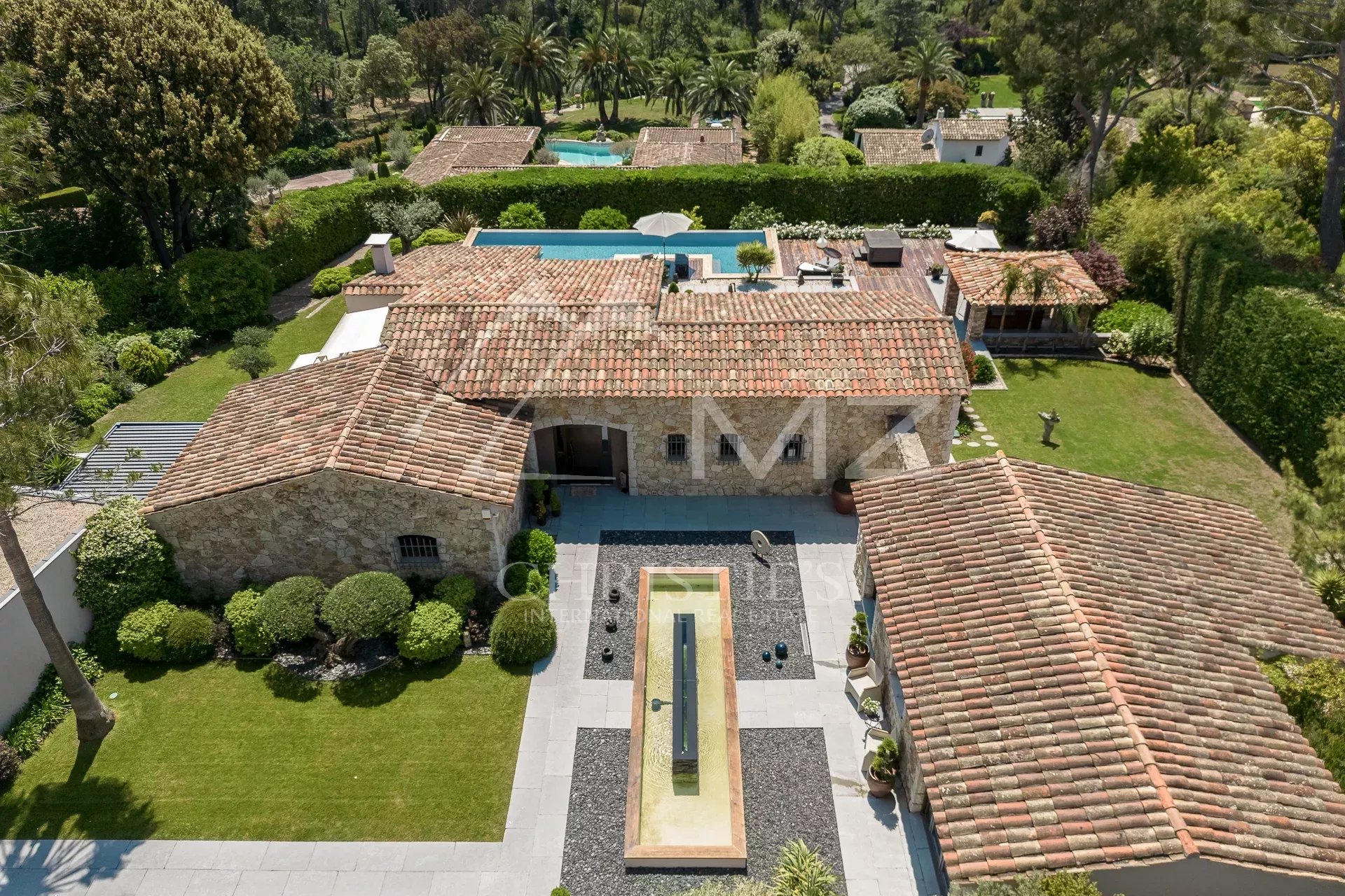 Aerial view of a stone villa with terracotta roofs, central courtyard, and a long rectangular water feature surrounded by gravel and greenery at the front.