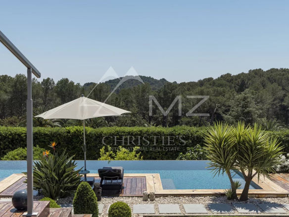 Infinity pool with a wooden deck, white umbrella and lounge chairs, surrounded by hedges and tropical plants with hills in the background.